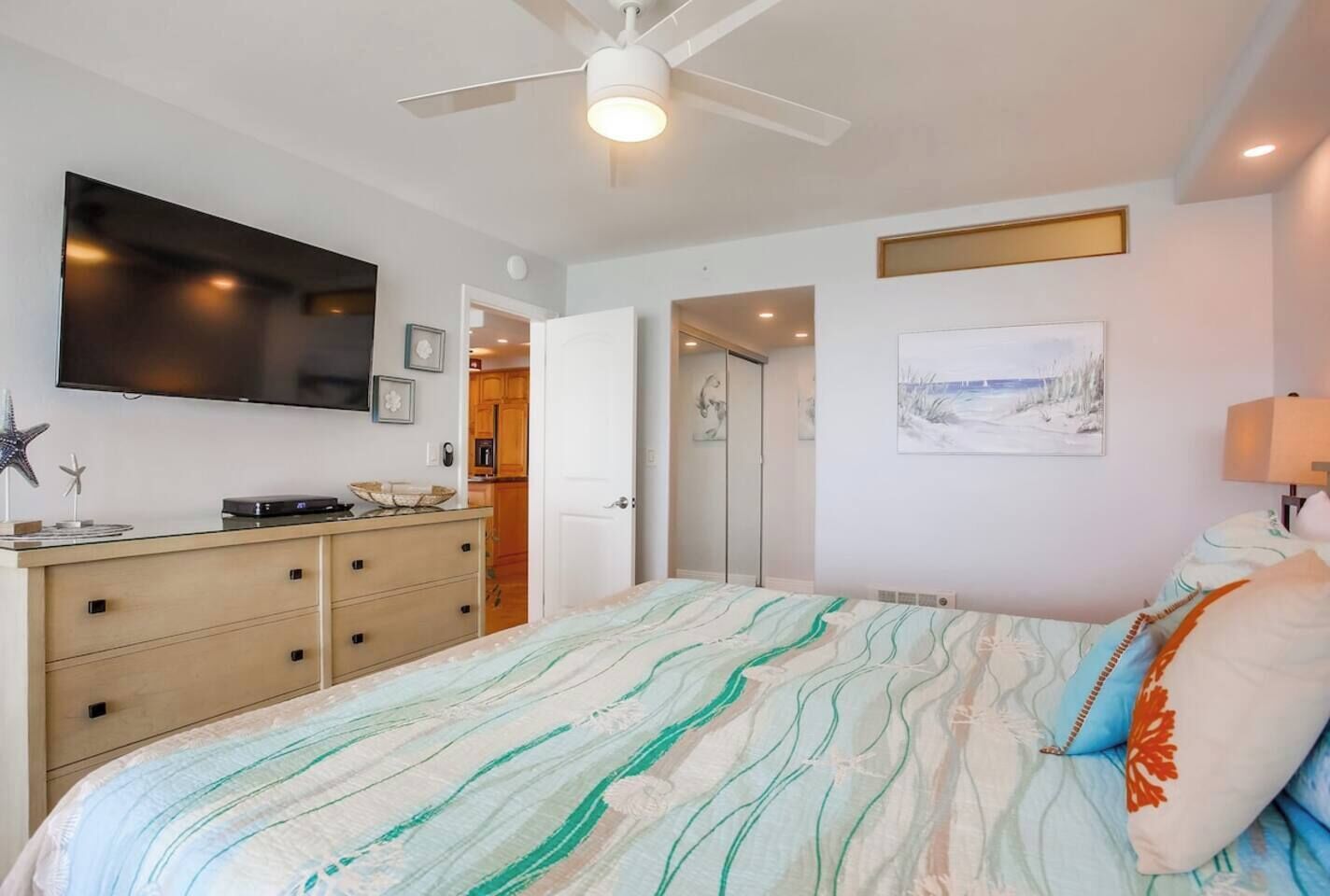 Een slaapkamer met een bed, een kaptafel, een televisie, een plafondventilator en een schilderij aan de muur.