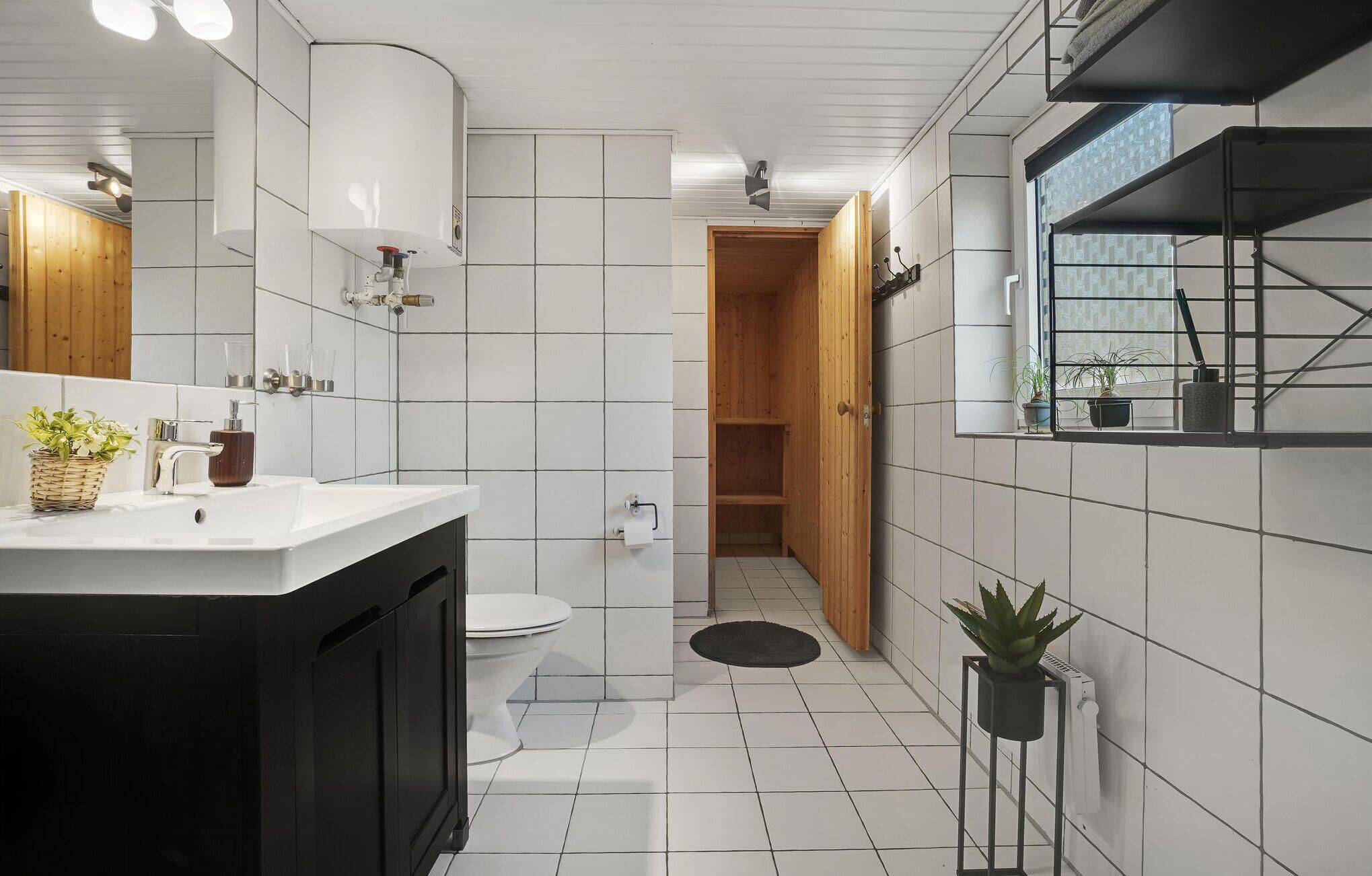 Un baño moderno con piso de baldosas blancas, un lavamanos negro con gabinete de madera, inodoro y cabina de ducha de madera.