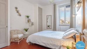 A bedroom with a large bed, a wicker chair, a small table with a plant, and a window with a view of buildings. - La Savoyarde - T2 apartment close to the lake. (Menthon-Saint-Bernard)