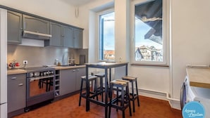 A modern kitchen with grey cabinets, a stove, and a window with a view of buildings. There are three stools around a table. - La Savoyarde - T2 apartment close to the lake. (Menthon-Saint-Bernard)