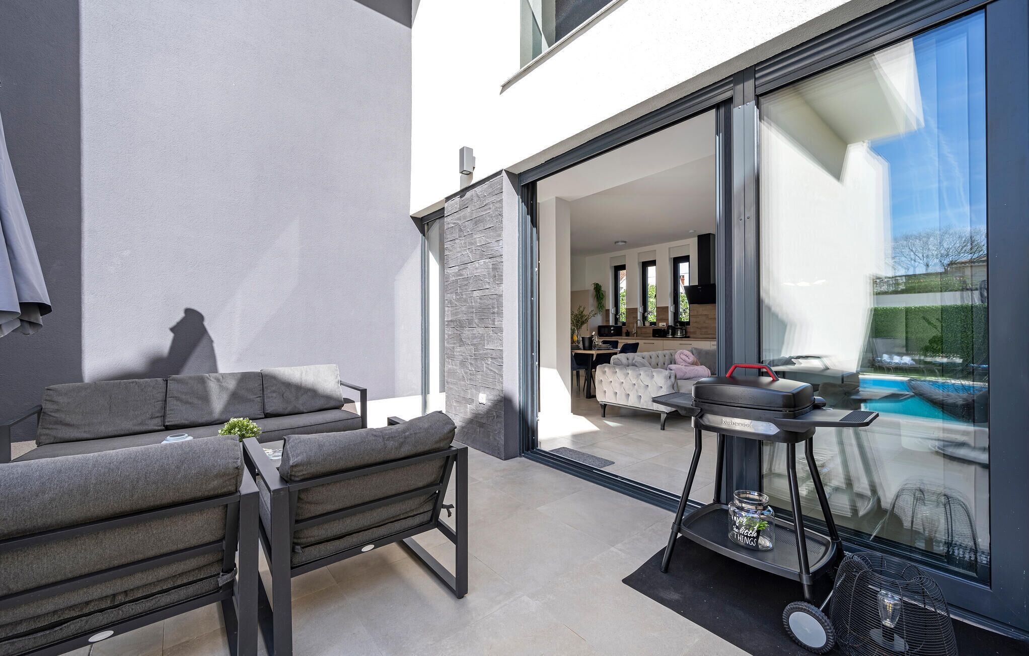 Un patio exterior con un sofá y una parrilla, una puerta de vidrio que da a una sala de estar, y una piscina visible a través del vidrio.