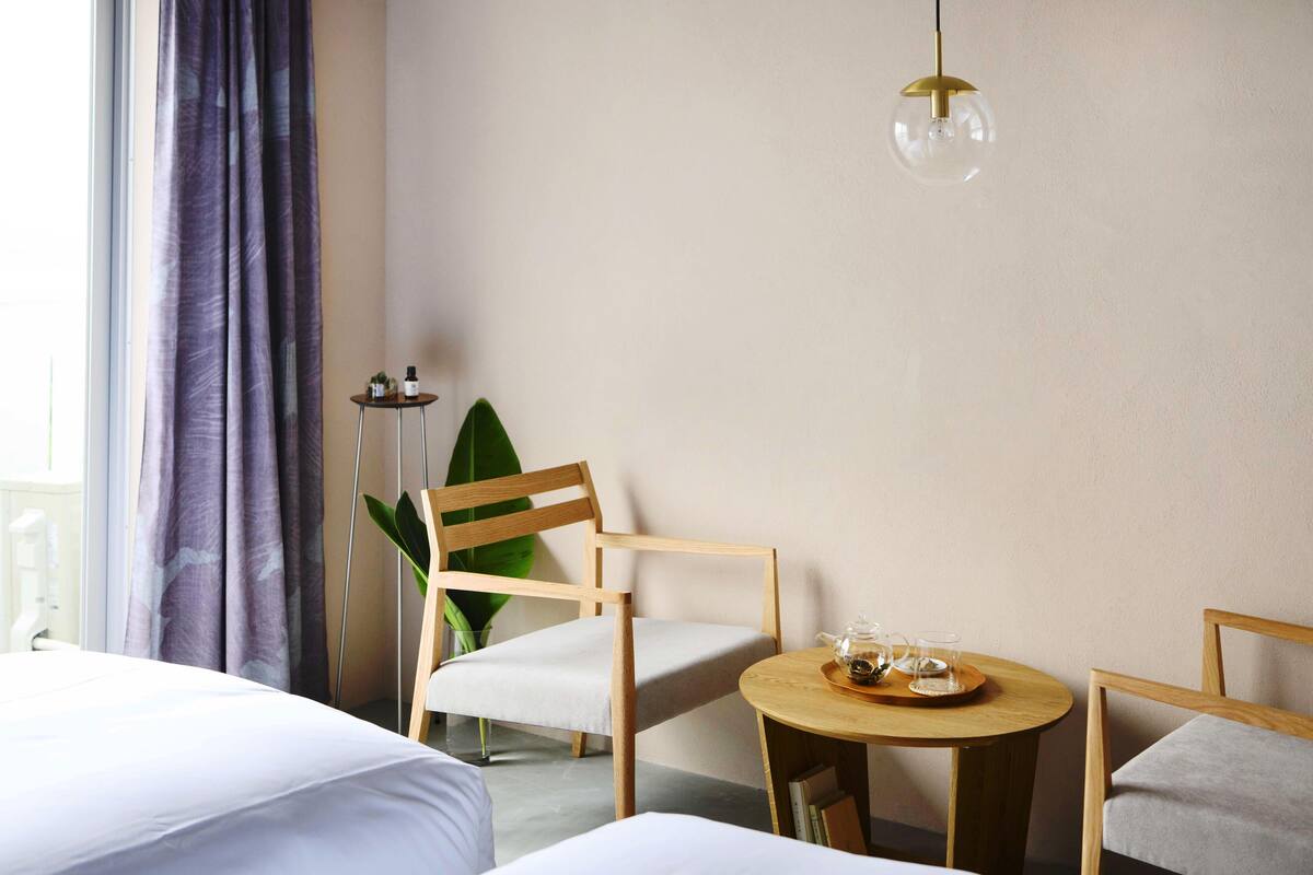 A hotel room with a bed, two chairs, a small round table with a tray, a potted plant, and a hanging light fixture.