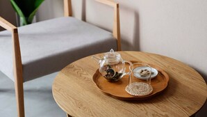 A round wooden table with a glass teapot and two glass cups on a woven coaster, set on a chair with a grey cushion. - Meguru Ishigaki (Ishigaki)