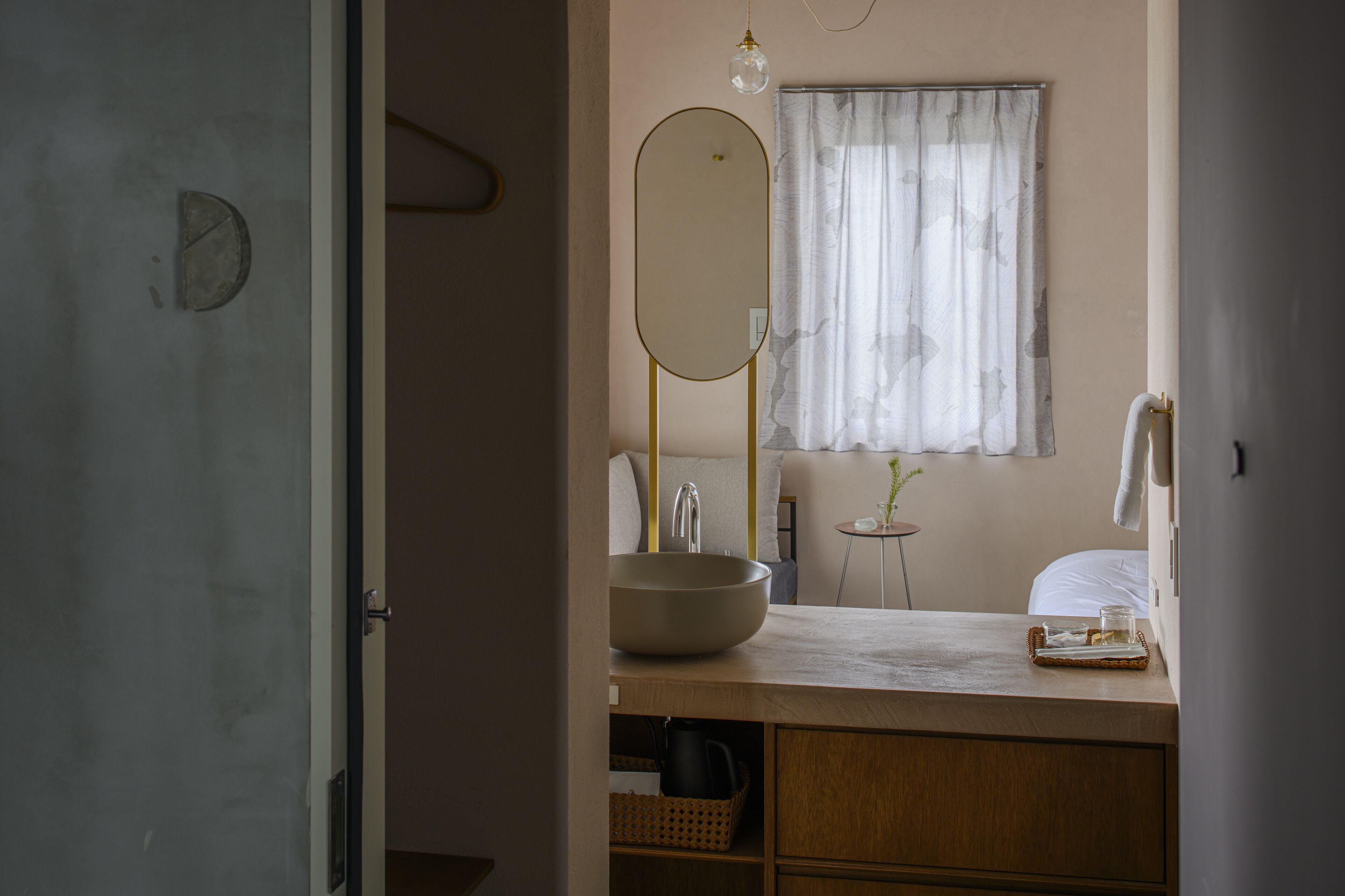 Un baño con un espejo ovalado grande, un lavabo redondo, una mesita con un vaso y una ventana con cortinas traslúcidas.