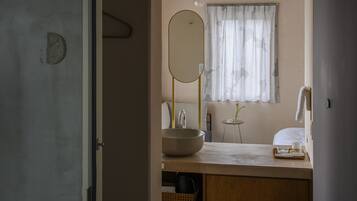 Un baño con un espejo ovalado grande, un lavabo redondo, una mesita con un vaso y una ventana con cortinas traslúcidas.