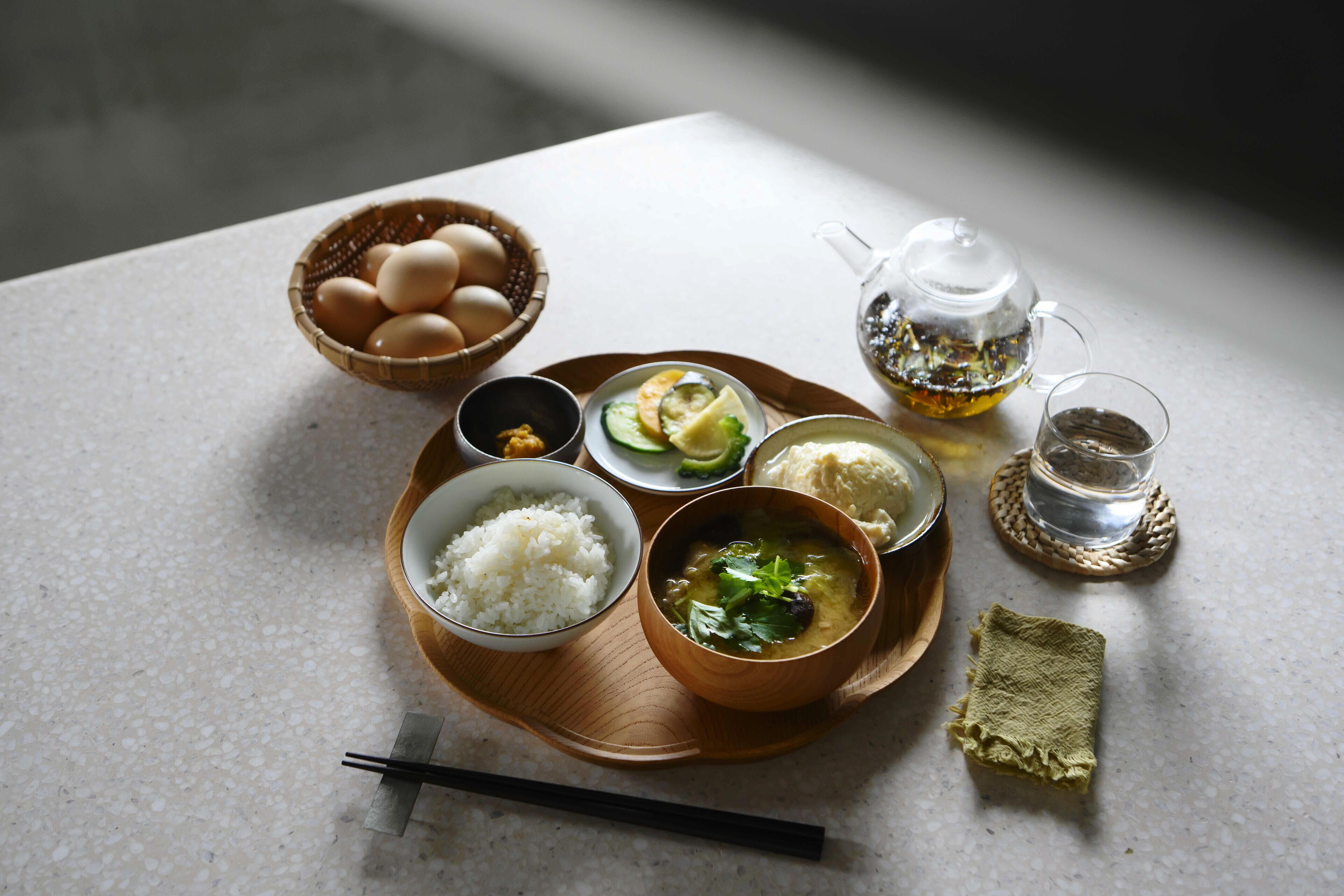 Una mesa con diversos alimentos, como arroz, huevos y sopa, junto con una tetera y un vaso de agua.