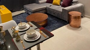 A modern living room with a grey sectional sofa, a round coffee table, and a dining area with a black table and gold cutlery. - Aminah’s Space - Jobz Luxury Rental (Bijilo)
