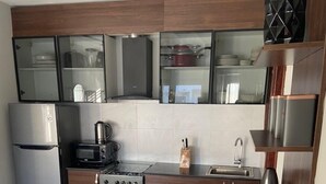 A modern kitchen with dark wood cabinets, a stainless steel refrigerator, a gas stove, and a black countertop. There are white chairs around a dining table set with white plates and gold utensils. - Aminah’s Space - Jobz Luxury Rental (Bijilo)