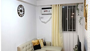 A living room with a beige sectional sofa, a small ottoman, a wall clock, an air conditioning unit, a window with curtains, and a television on a stand. - Modern minimalist condo in Davao City & very near to Samal Island (Pet Friendly) (Davao City)