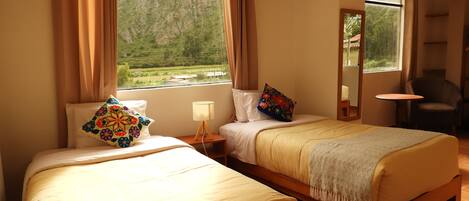Habitación de hotel con dos camas, una mesita de noche con lámpara, una silla y vistas a un paisaje verde a través de la ventana.