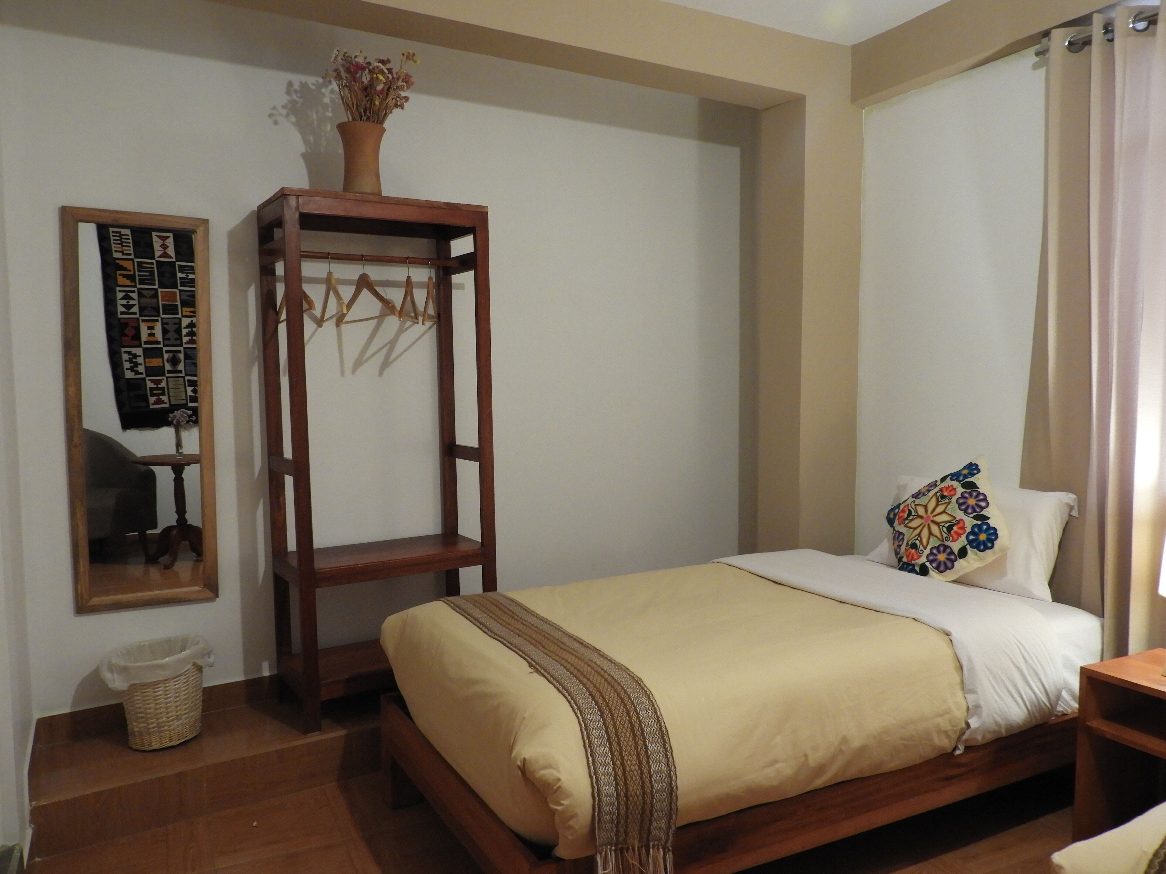 A bedroom with a wooden bed, a small wooden shelf with a vase, a wall-mounted quilt, and a wicker basket.