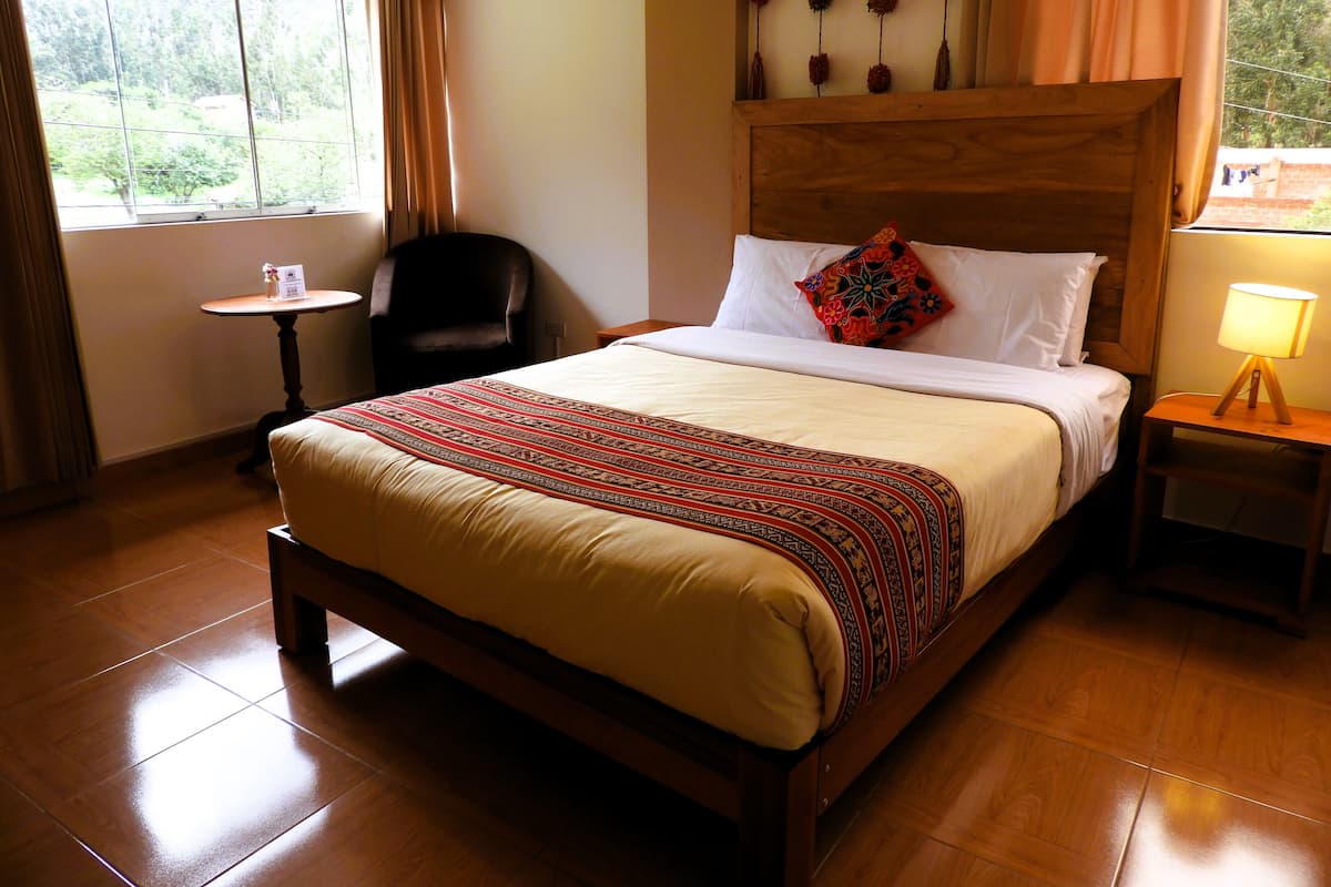 A hotel room with a wooden bed, a small table, a chair, and a window with curtains.