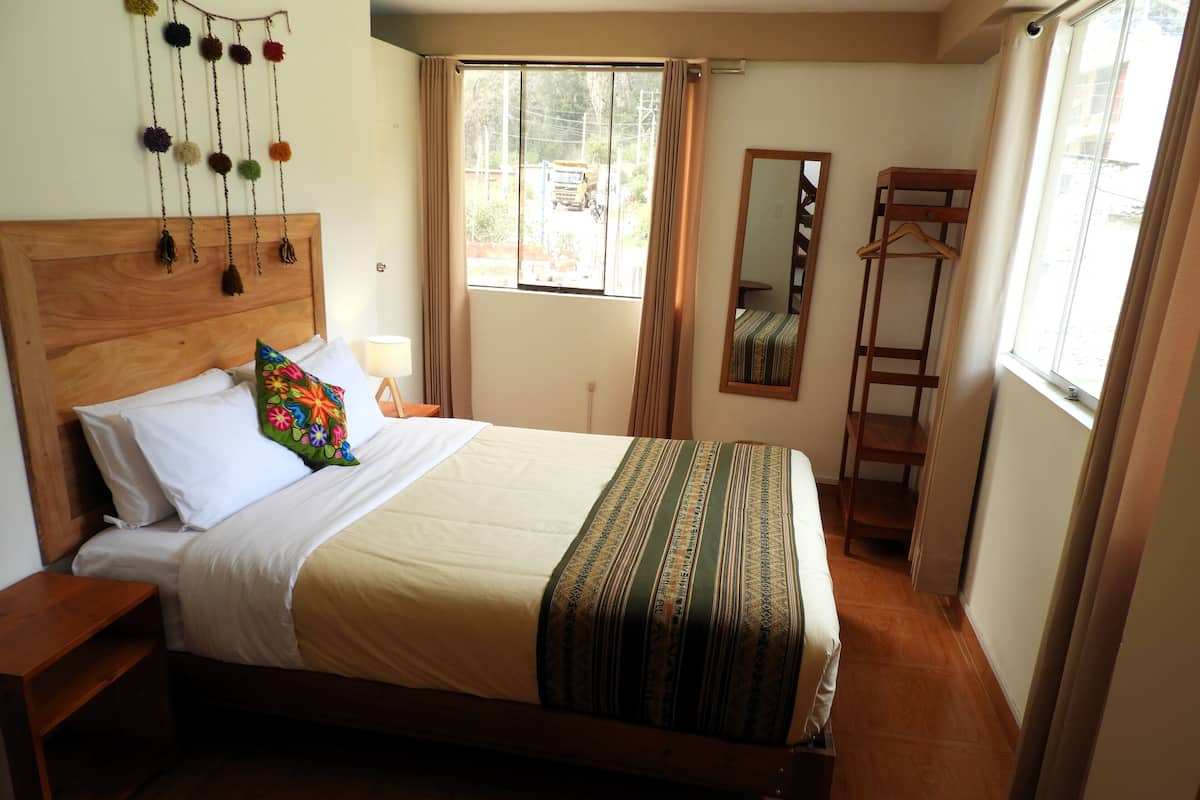 A neatly made bed with a wooden headboard, a colorful pillow, and a striped blanket. There is a wooden nightstand with a lamp, a window with curtains, and a wooden ladder against the wall.