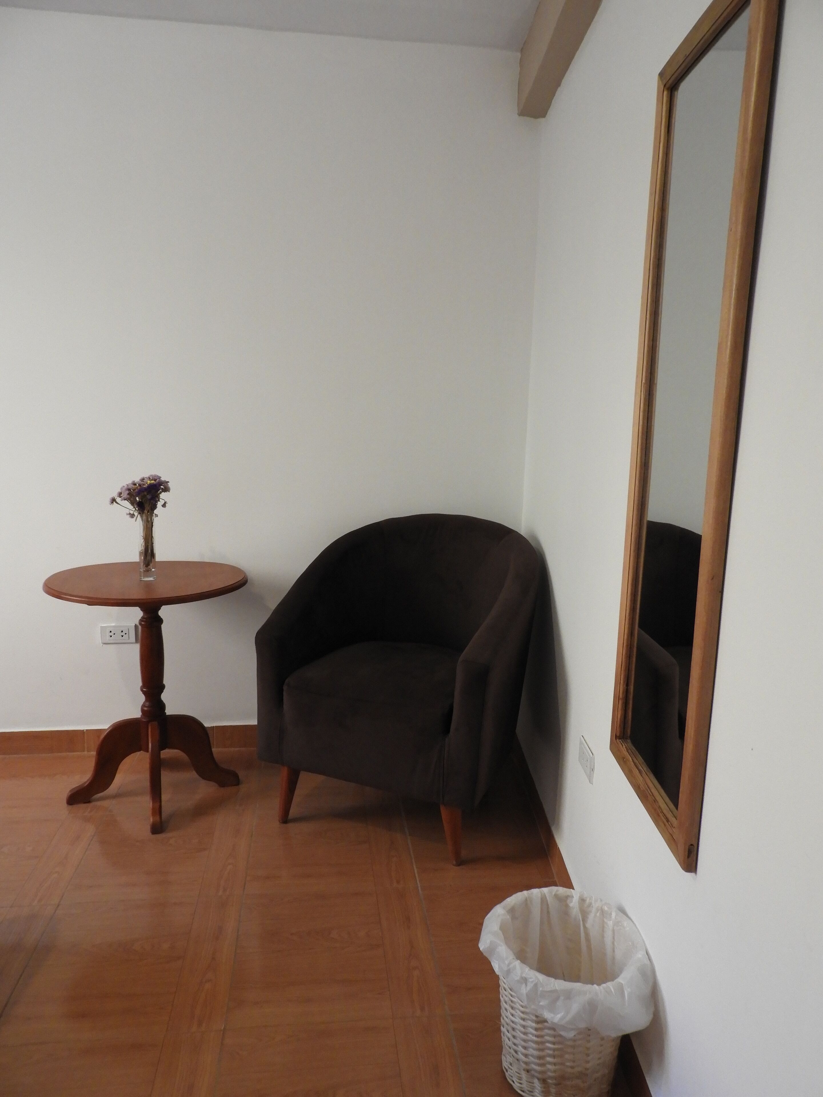 A room with a wooden floor, a dark armchair, a small round table with a vase, and a large mirror on the wall.