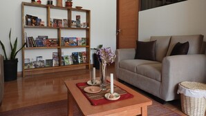Un salón con sofá, una mesa de centro de madera con objetos decorativos, una estantería con libros y adornos, y un ventanal con persianas.