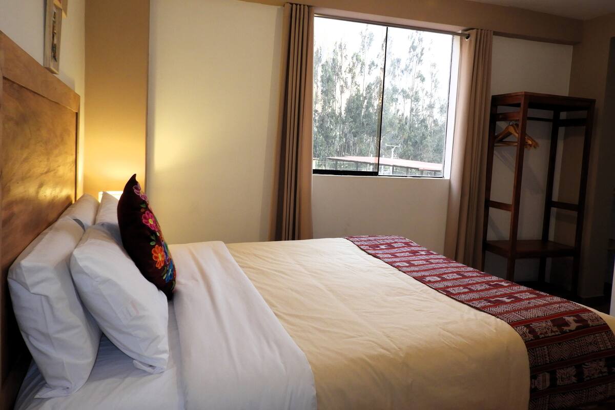 A hotel room with a bed, a window with curtains, a wooden headboard, and a wooden wardrobe.