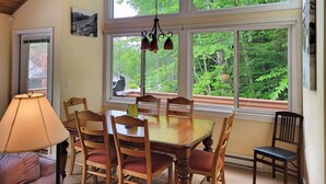 Zona pranzo con tavolo e sedie in legno, una lampada e ampie finestre con vista su piante.