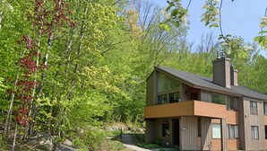 Una moderna casa a due piani con un ampio balcone, immersa in un paesaggio naturale tra alberi e rocce.