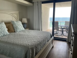 A hotel room with a bed, a nightstand, a lamp, a view of the ocean through a balcony door, and a table and chairs outside.