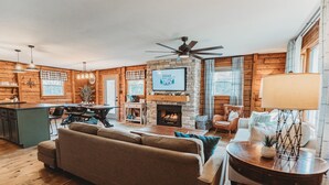 A cozy living room with a fireplace, a television mounted above it, and a dining area with a kitchen in the background. - Fire Pit, 10 private Acres | Pets Welcome (Bourbon)