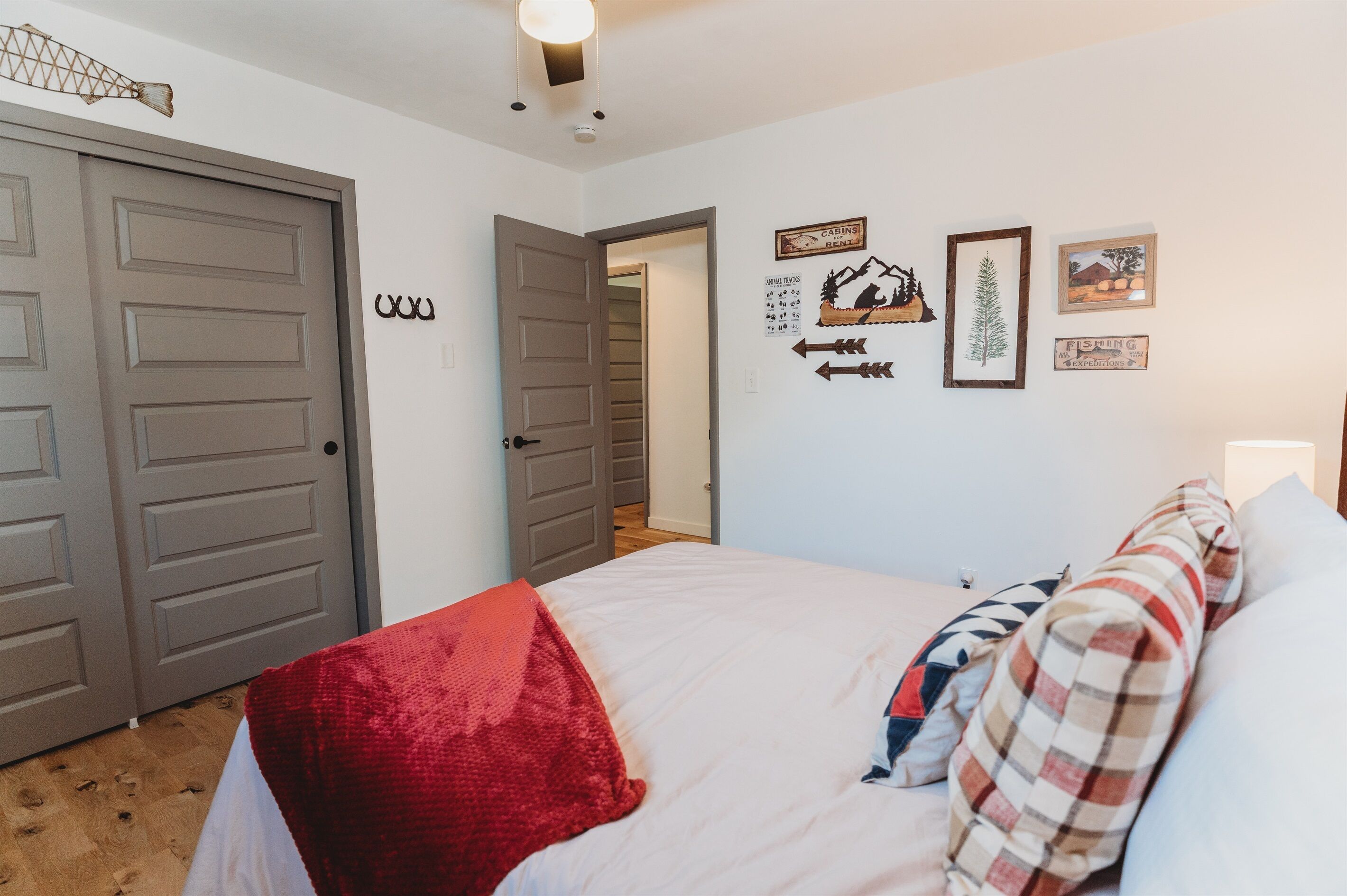 Une chambre à coucher avec un lit, des oreillers, un plaid rouge et une porte donnant sur une autre pièce. Les murs sont décorés, notamment avec un tableau encadré représentant une montagne et un panneau indiquant « CABANES ».