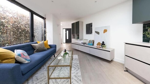 A modern living room with a blue sofa, a glass coffee table, and a view of the outdoors through large windows. - Cozy London House with Garden & Terrace | Near Westfield Stratford & Tube (London)
