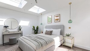A modern bedroom with a skylight, a bed with a grey blanket, a desk with a mirror, and a small side table with a plant. - Cozy London House with Garden & Terrace | Near Westfield Stratford & Tube (London)