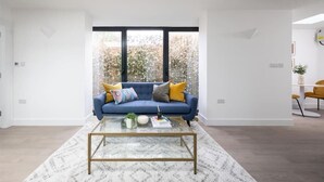 A modern living room with a blue sofa, a glass coffee table, and a large window with a view of trees. - Cozy London House with Garden & Terrace | Near Westfield Stratford & Tube (London)