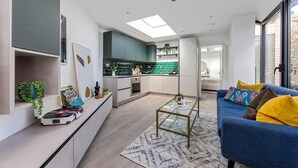 A modern kitchen with a central island, a dining area with a blue sofa, and a large window providing natural light. - Cozy London House with Garden & Terrace | Near Westfield Stratford & Tube (London)