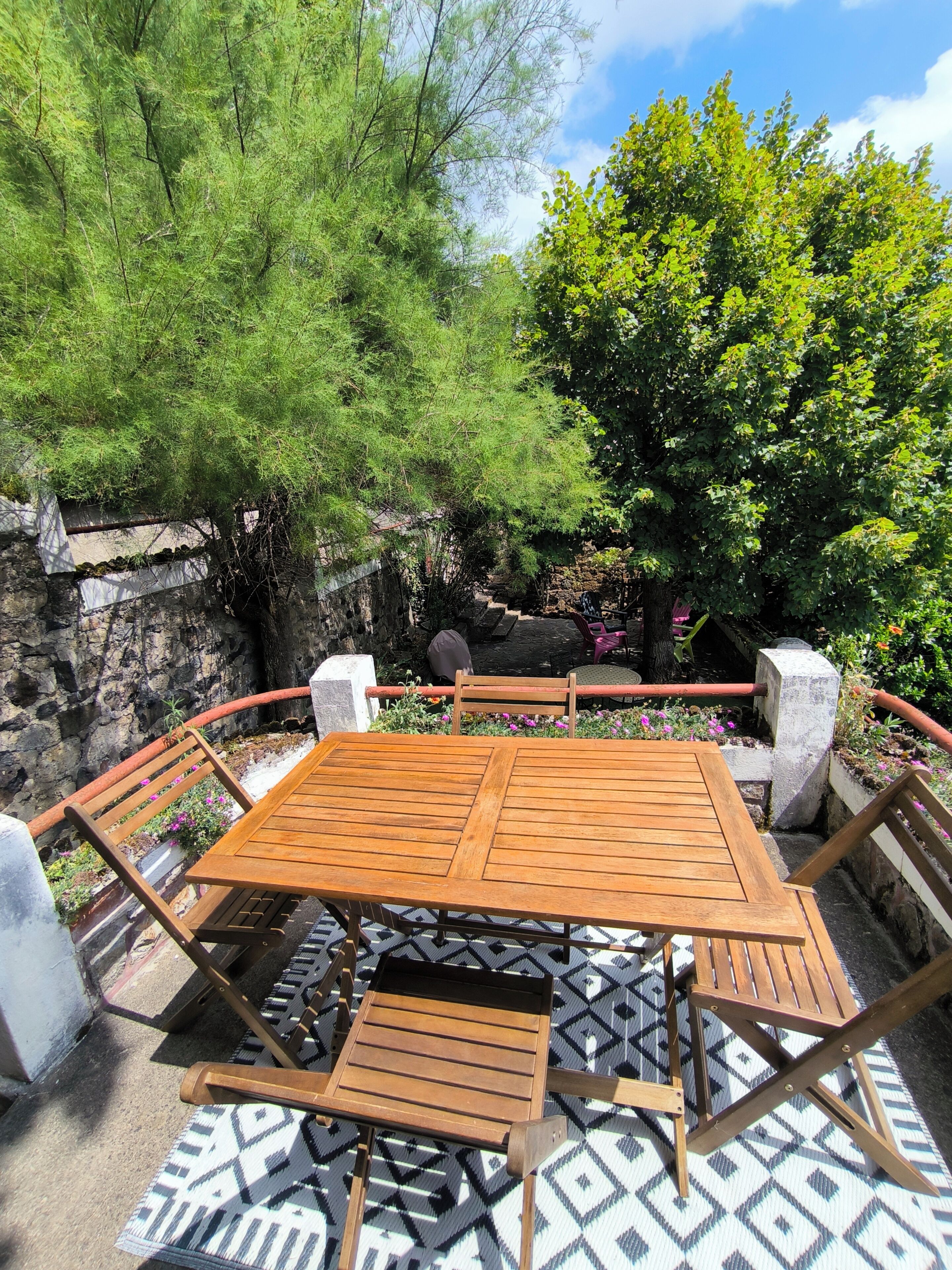 An outdoor seating area with a wooden table and chairs, surrounded by greenery and a stone wall.