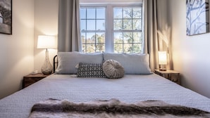 A neatly made bed with a white quilt, two pillows, and a fur throw. There are two bedside lamps on wooden nightstands. A window with curtains is visible, and a painting hangs on the wall. - Tranquil Lakefront Farm Oasis ~ Pool ~ Scenic View<br>  (Bumpass)