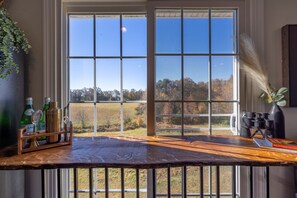 A wooden bar counter with a view of a field and trees through large windows. - Tranquil Lakefront Farm Oasis ~ Pool ~ Scenic View<br>  (Bumpass)