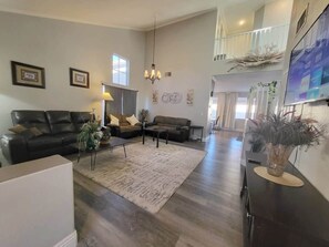 Ein geräumiges Wohnzimmer mit einer großen Couch, einem Couchtisch und einem Teppich. An der Wand befinden sich zwei gerahmte Bilder und eine Chandelieber hängt von der Decke.