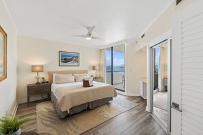 A bedroom with a bed, bedside tables, a ceiling fan, a large window with a city view, and a sliding glass door leading to a balcony. - Beachfront at Destin Harbor's East Pass! Epic Views, Pool, Private Beach! (Destin)