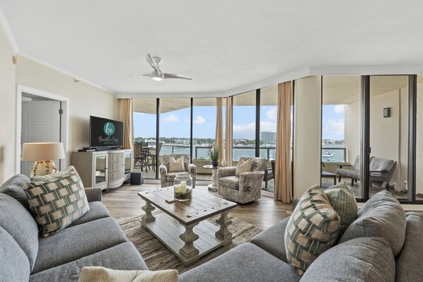 A spacious living room with a large sofa, a coffee table, and a TV. There are two armchairs and a dining table with chairs. The room has large windows offering a view of the water and buildings. - Beachfront at Destin Harbor's East Pass! Epic Views, Pool, Private Beach! (Destin)