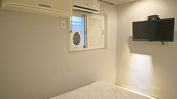 A small, single-bed room with a window, an air conditioning unit, and a mounted television.