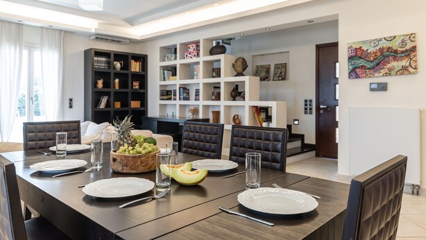 Een moderne eethoek met een donker houten tafel voor vier personen, omringd door donker lederen stoelen. Op tafel staat een fruitschaal met druiven en een meloen.