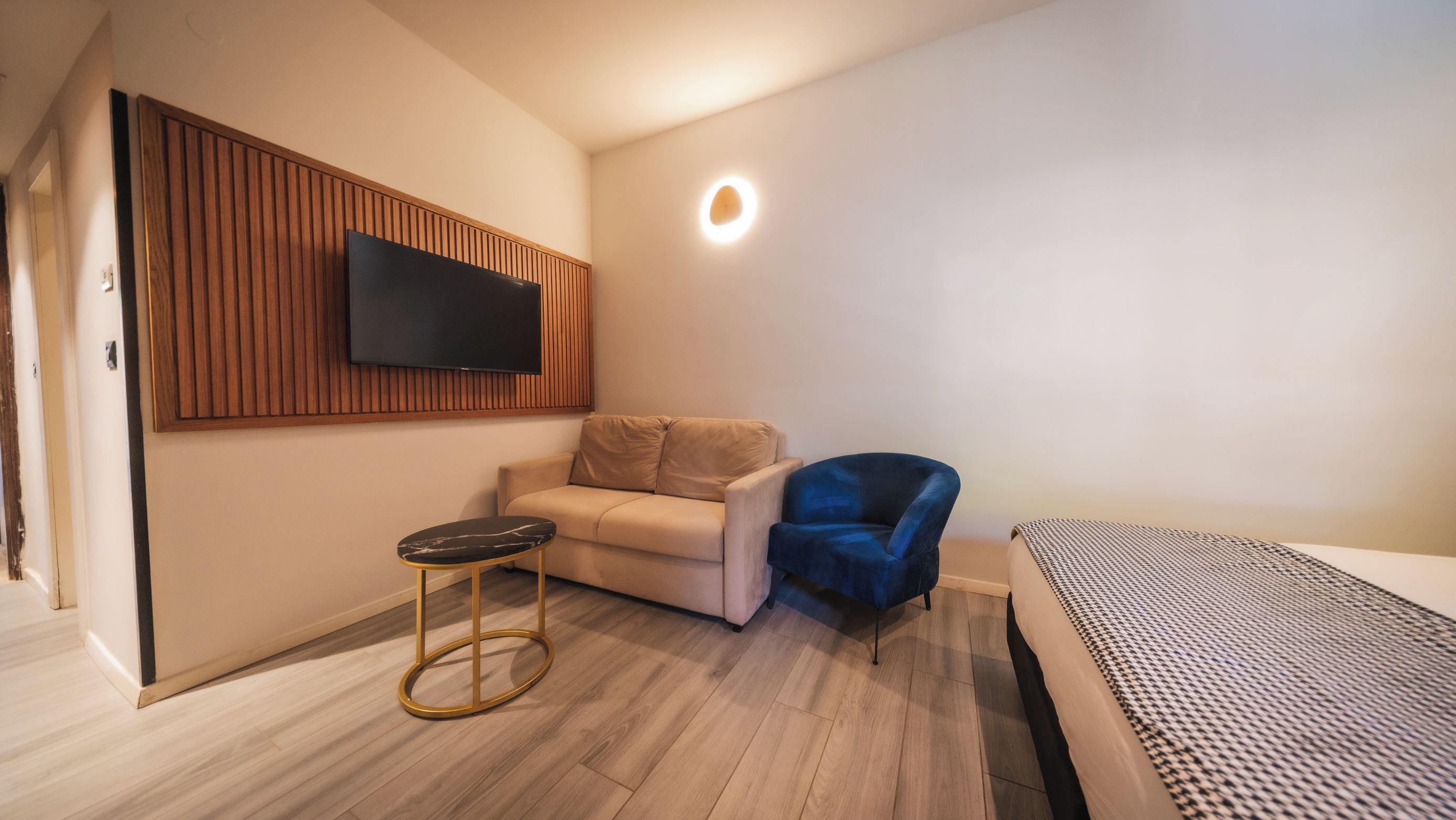A modern room with a flat-screen TV mounted on a wooden panel wall, a beige sofa, a blue chair, a small round table, and a bed with a patterned bedspread.