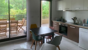 Et moderne køkken med hvide skabe, en indbygget ovn og en opvaskemaskine. Der er et spiseområde med et bord og stole, samt en balkon med udsigt over grønne områder.