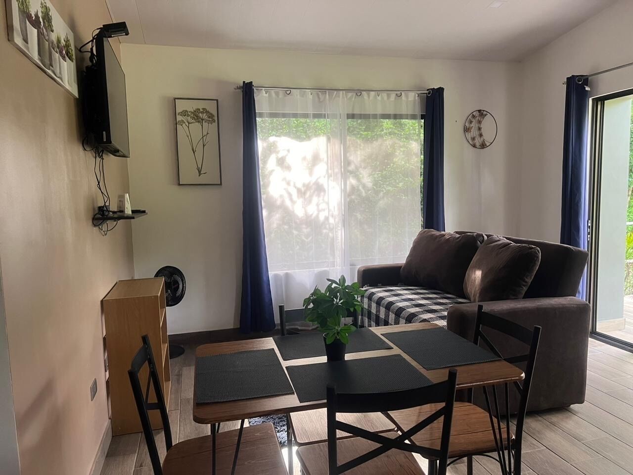 Un soggiorno con un divano, un tavolo da pranzo con sedie, una piccola pianta e una TV montata a parete.
