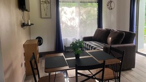 Una sala de estar con un sofá, una mesa de comedor con sillas, una planta pequeña y un televisor montado en la pared.