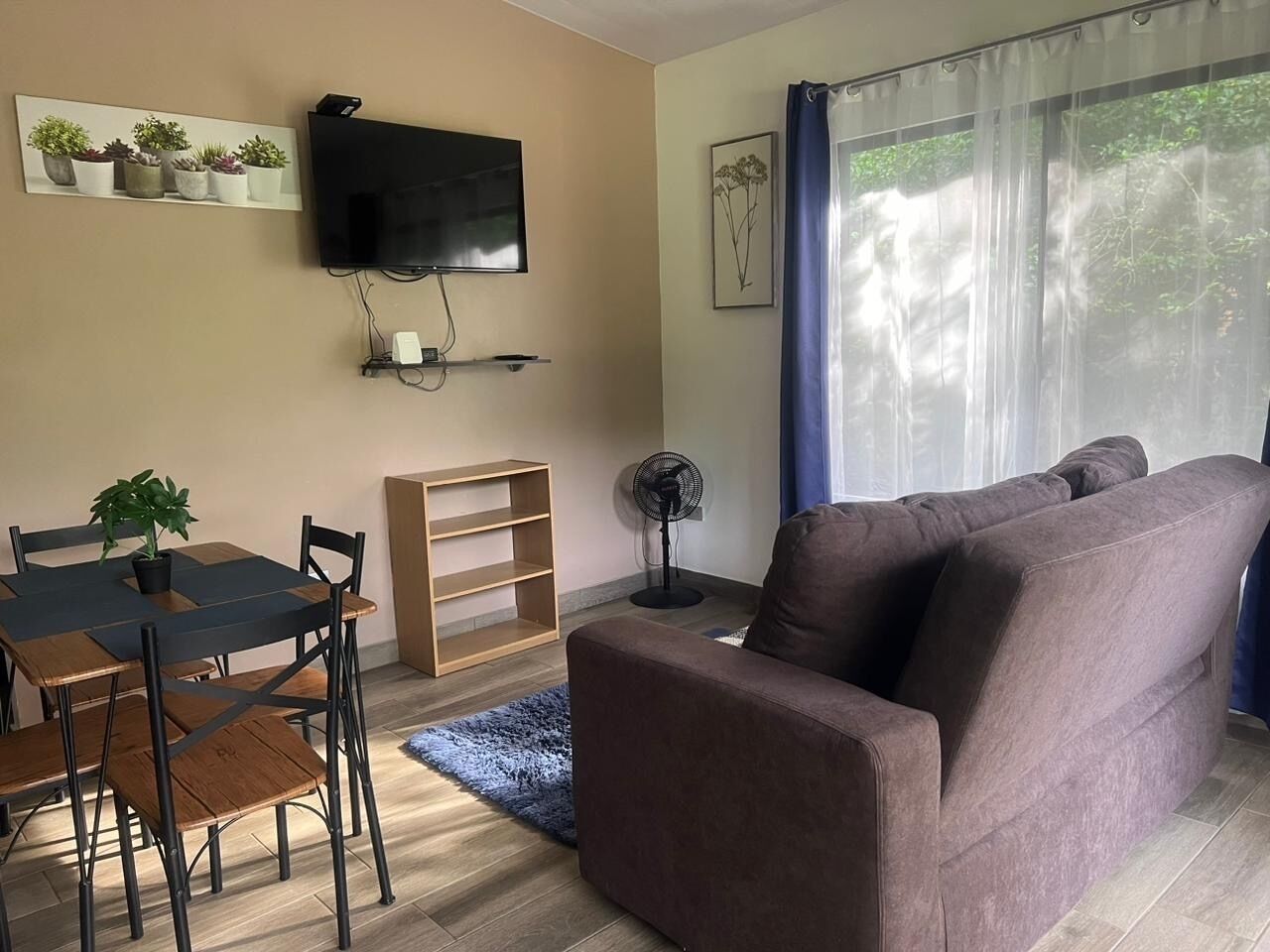 A living room with a brown sofa, a dining table with chairs, a TV mounted on the wall, and a fan on the floor.