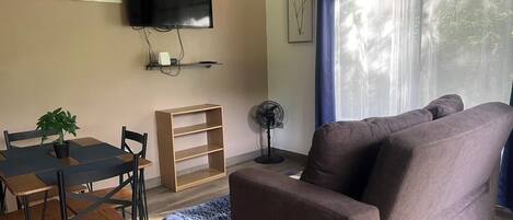 A living room with a brown sofa, a dining table with chairs, a TV mounted on the wall, and a fan on the floor.