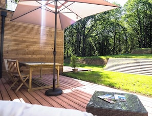 Une terrasse en bois avec une table et des chaises, un grand parasol et une table en verre avec des magazines.
