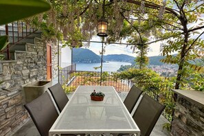 Um terraço com mesa e cadeiras, um candeeiro pendurado e vista para um lago e montanhas.