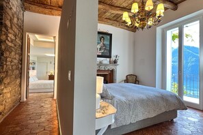 Quarto com cama, lustre, lareira, janela ampla e vista, e parede de pedra.