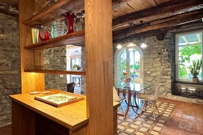 A rustic interior with a wooden table displaying a framed botanical print, a stone wall, and a view of greenery outside.