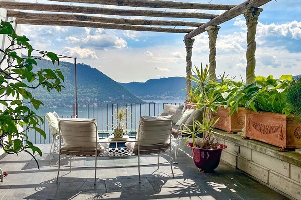 A terrace with a table and chairs, potted plants, and a view of a lake and mountains.