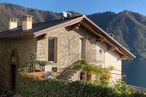 Uma casa de pedra com varanda, uma escadaria de madeira e vista para um lago e montanhas.