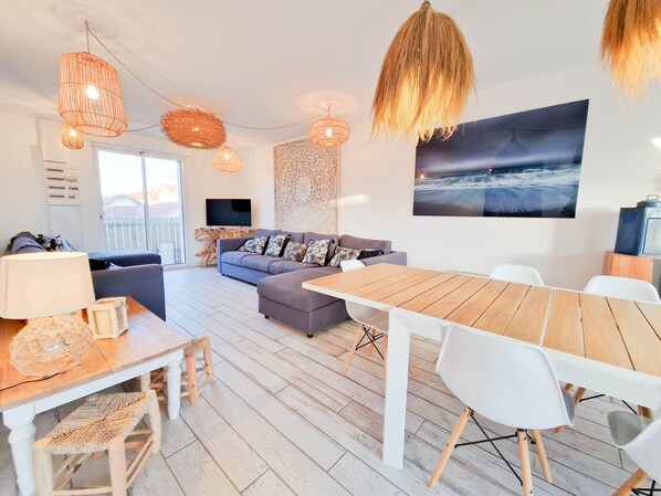 Ett modernt vardagsrum med en grå soffa, ett matbord i trä och vita stolar. En tv är monterad på väggen och ett stort fönster ger utsikt över omgivningen.